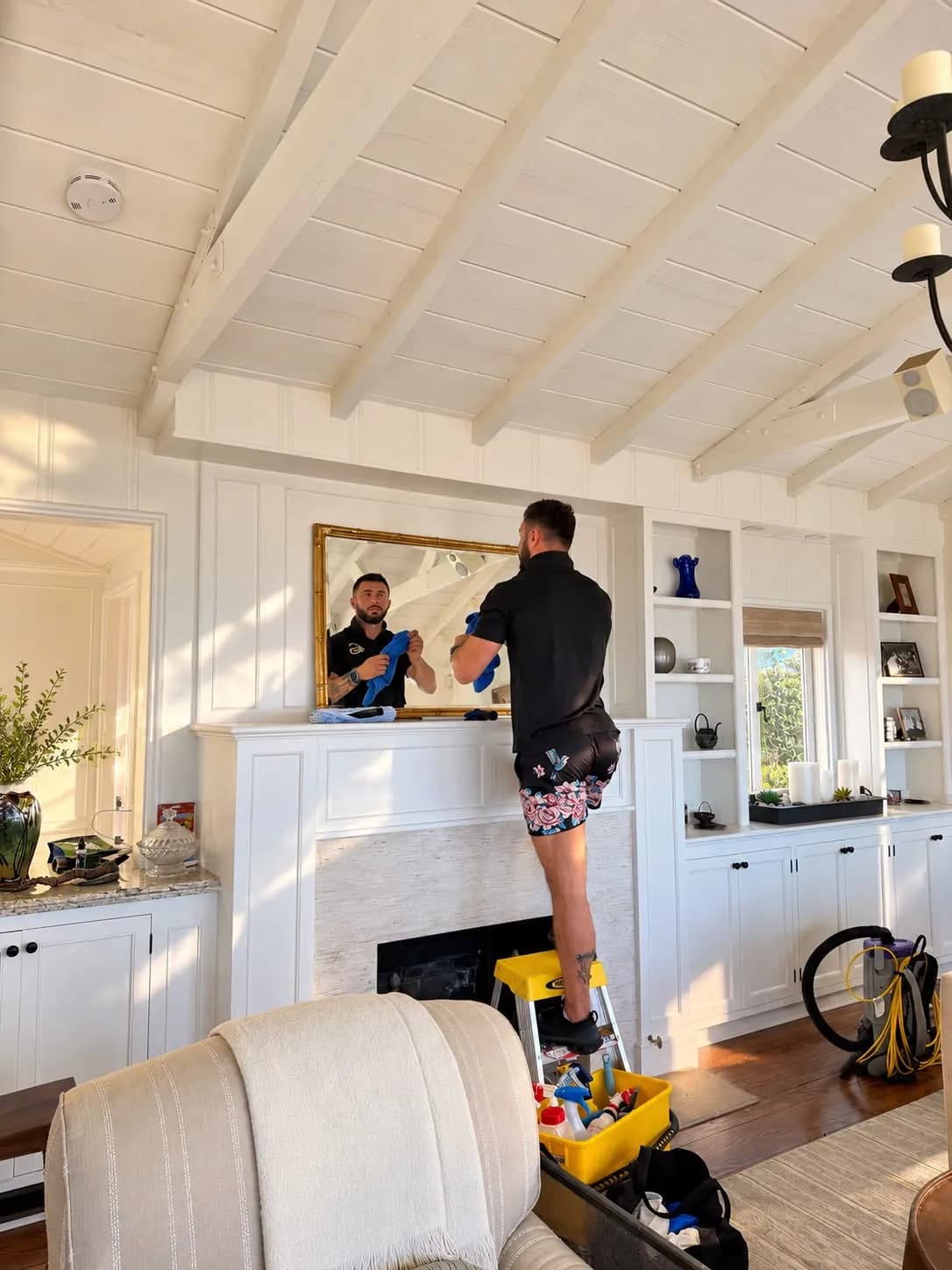 Polishing a gold-framed mirror above a white fireplace mantel